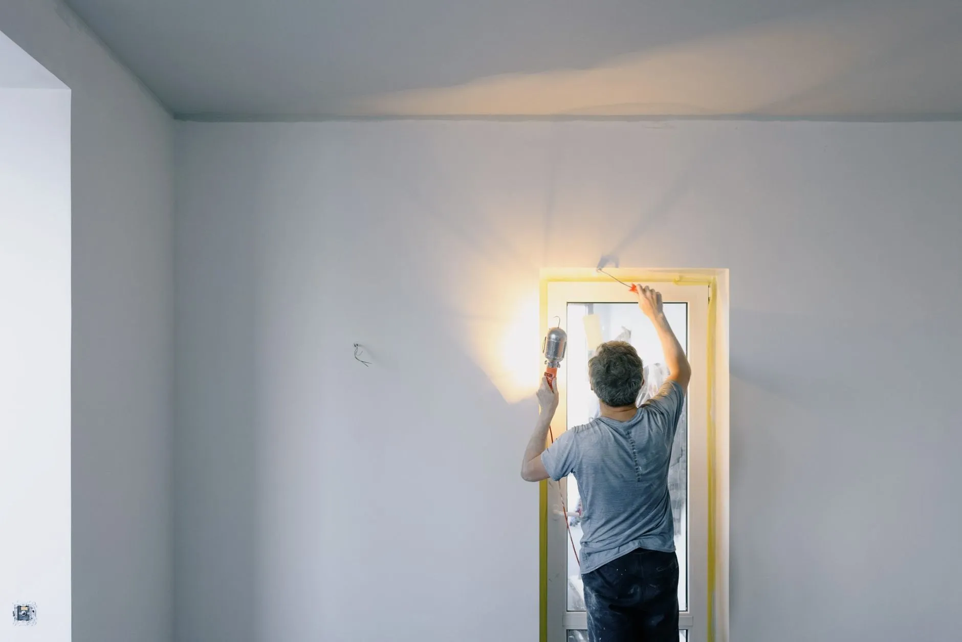 A decorator painting the top of a door frame by the light of a work lamp in a pale grey room