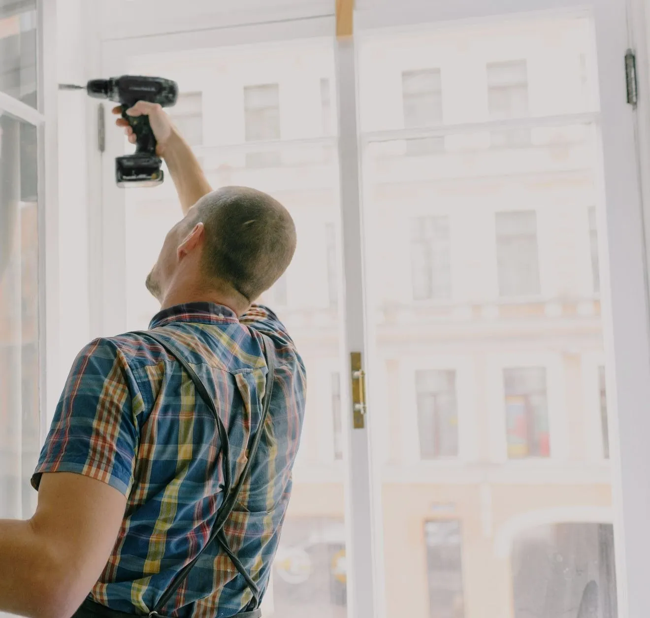 A handyman in a checked shirt driving a screw into the hinge of a tall open sash window