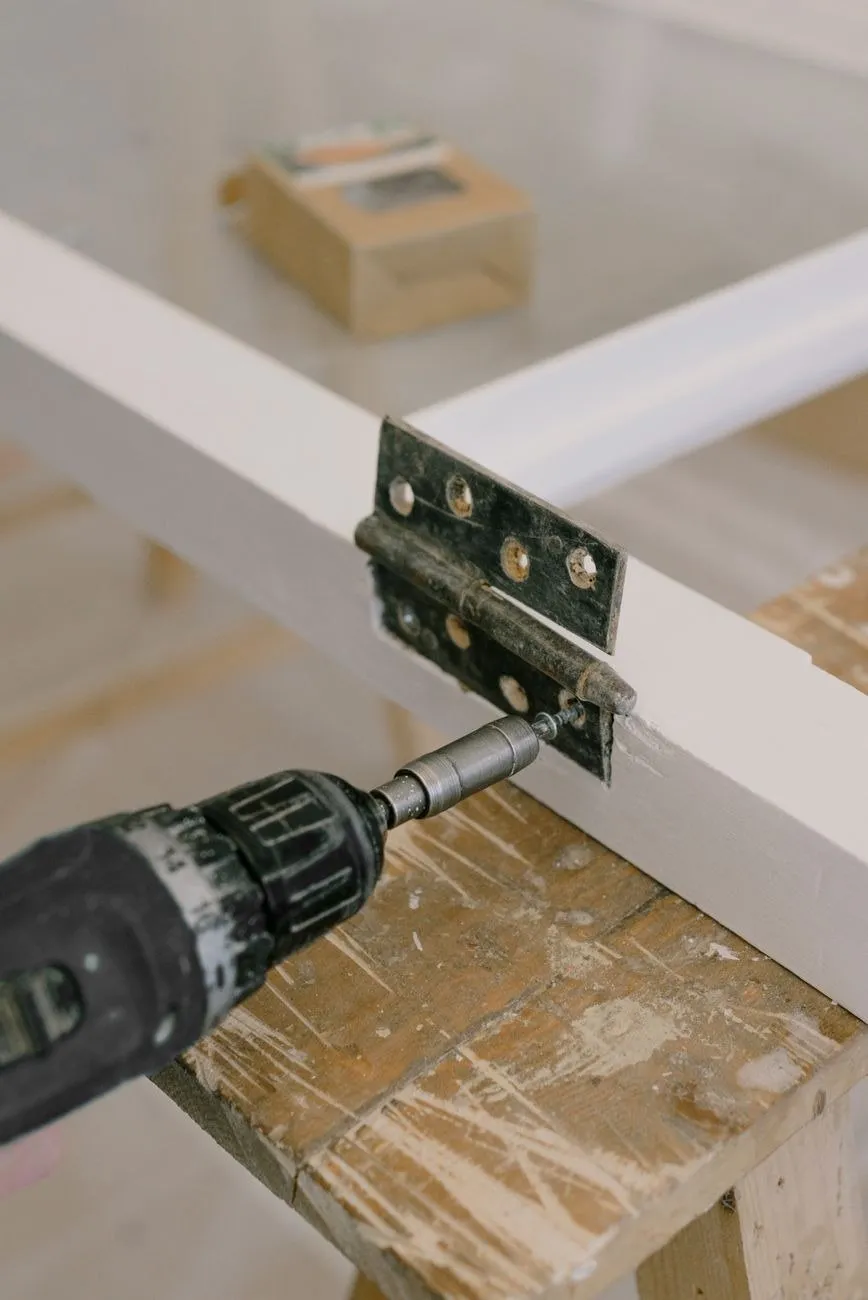 A hinge being screwed to the edge of a white painted door resting on a worn trestle