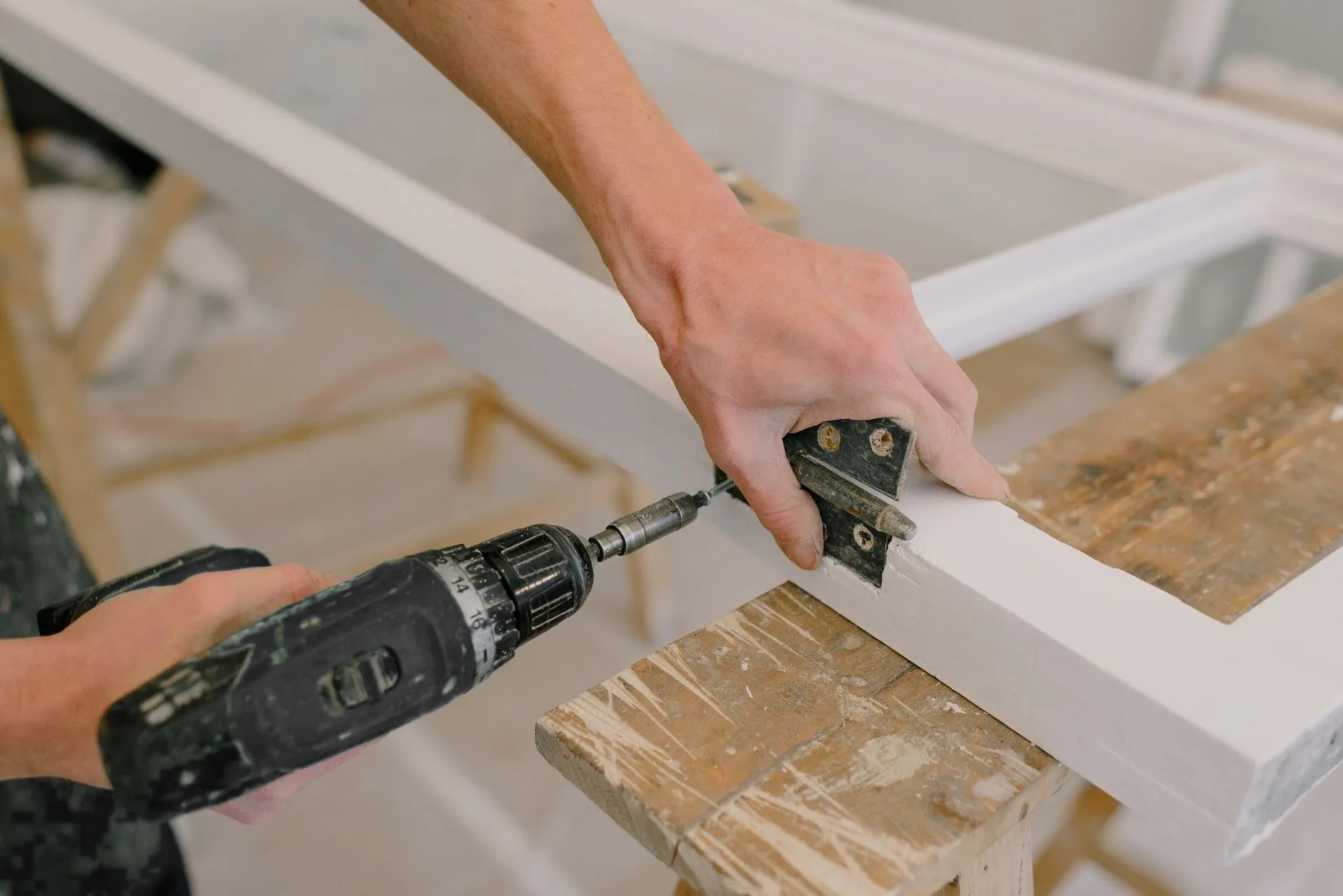 A driver turning a screw into a black hinge on the edge of a white frame resting on a trestle