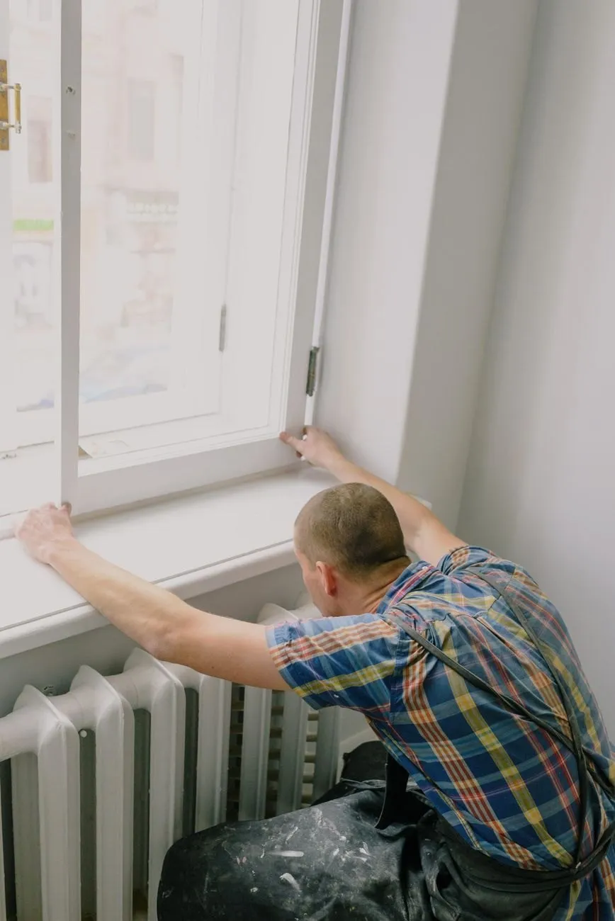 A handyman in a checked shirt adjusting a white timber window above a cast-iron radiator