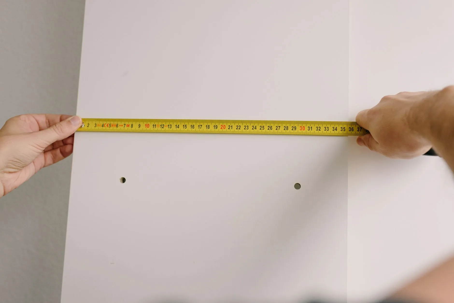 Two hands holding a tape measure across a wall between two drilled holes