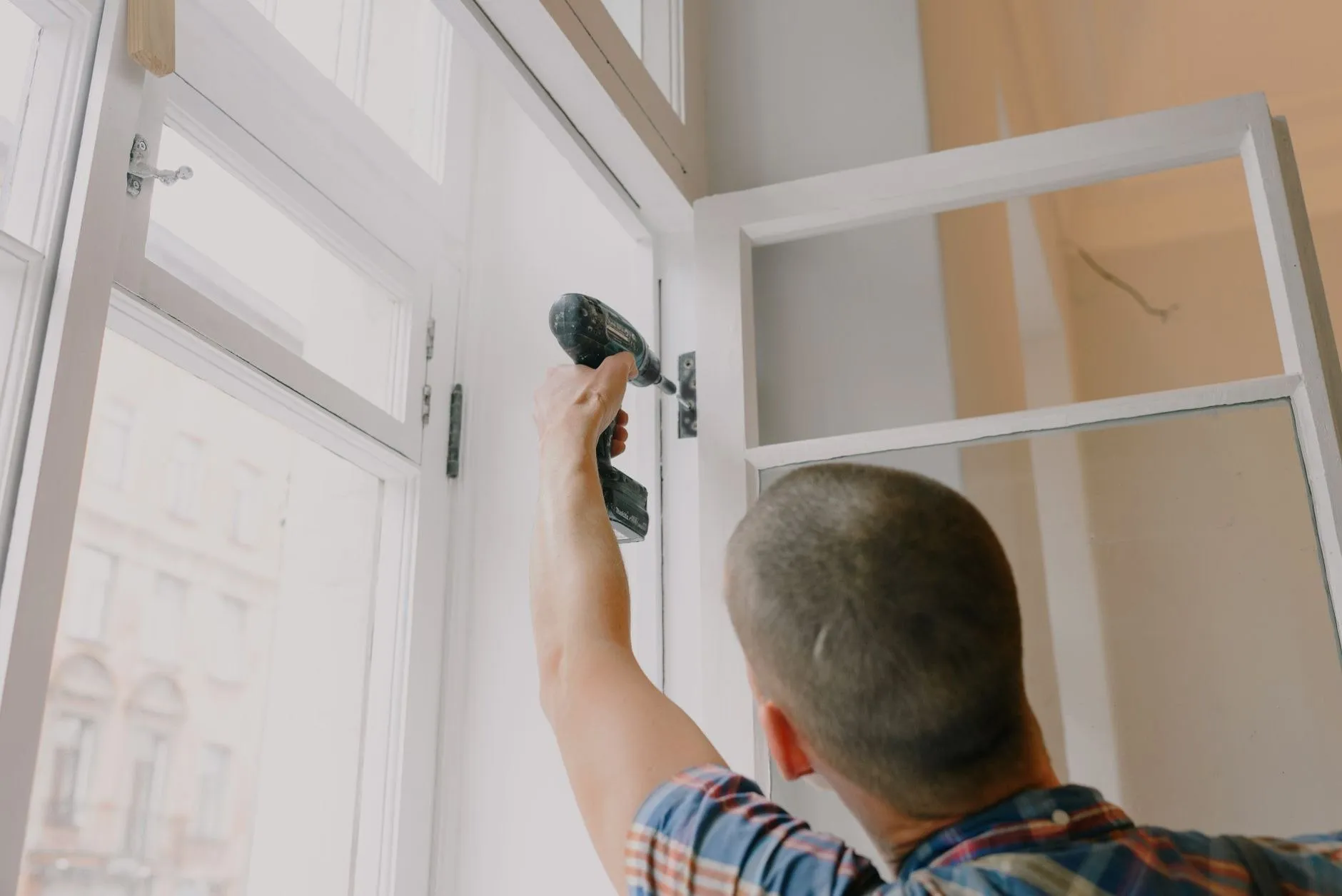 A handyman reaching up to drive a screw into the hinge of an open white window frame
